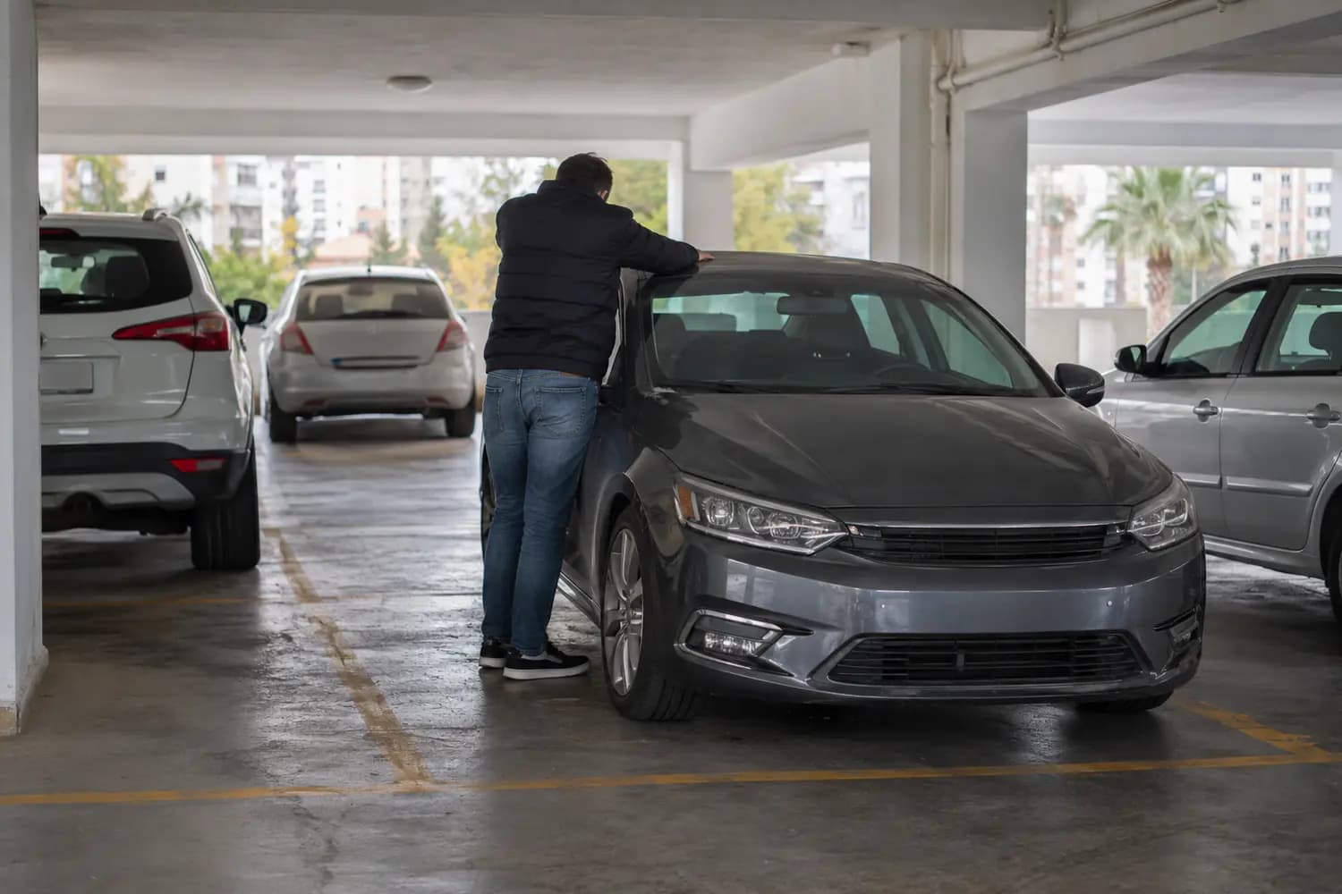 Apartman otoparkında çalışmayan, kaputu kapalı bir otomobil ve yanında duran sürücünün elleri